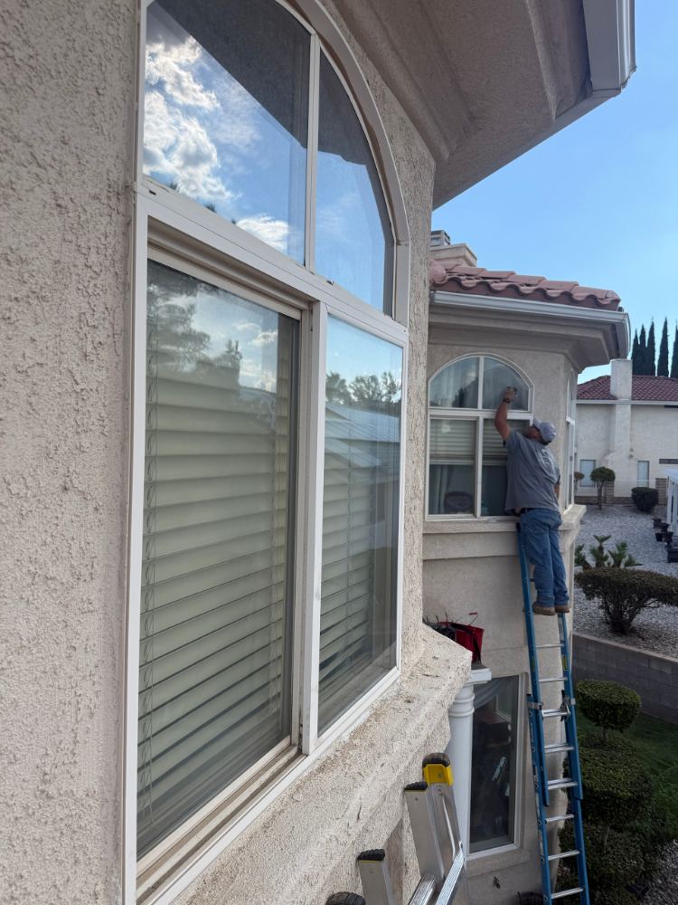 Two story stucco home in Spring Valley Lake with tall arched windows, technician on a ladder cleaning the upper glass