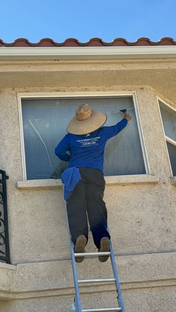 Technician in a Tony's Window Cleaning shirt squeegeeing a second story window from a ladder in Spring Valley Lake